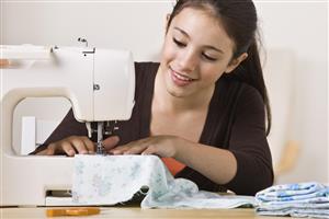 Young woman using a sewing machine with a stack of fabric nearby.